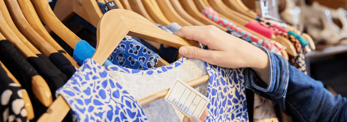 a rail of second hand clothes in many colours, a hand has pulled out a hanger with a blue and white patterned top on it.