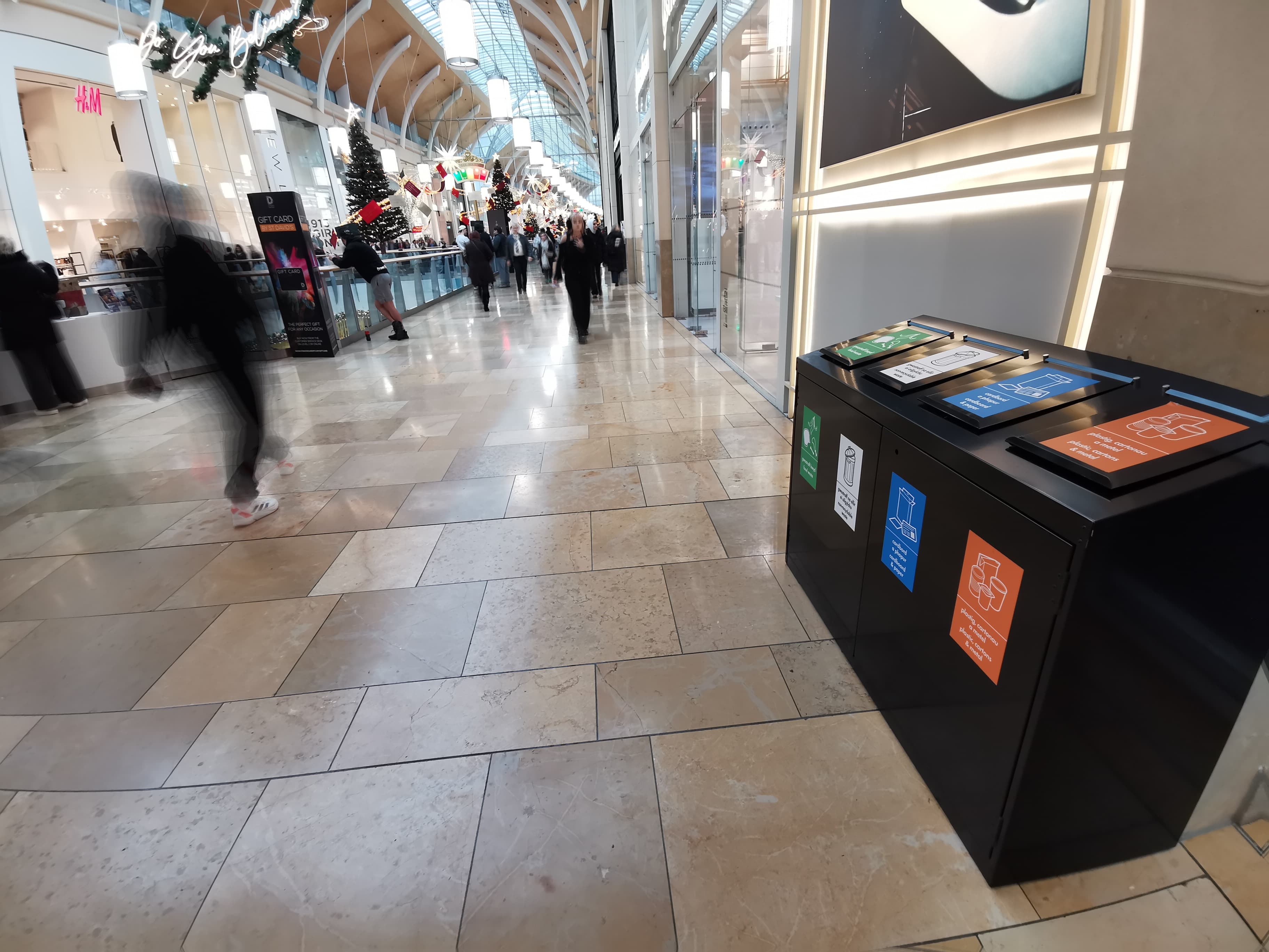 Rhes o finiau ailgylchu du gyda chaeadau o wahanol liwiau mewn canolfan siopa