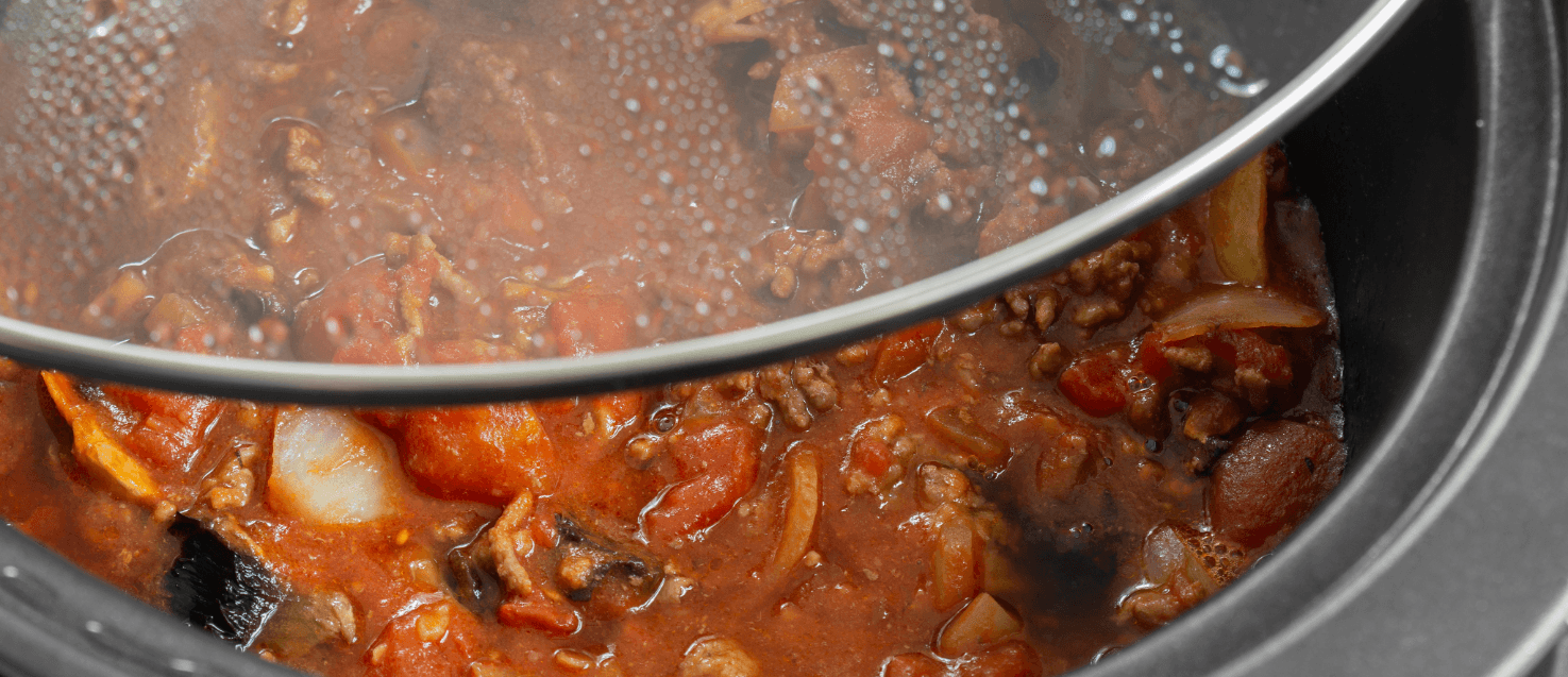 close-up of a slow cooker with tasty mince ina  tomato sauce withonions and over veggies and herbs